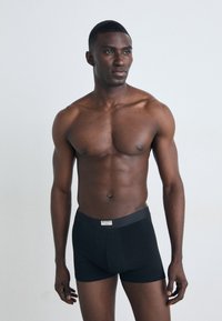 Zwarte stoffen boxershorts met een gladde textuur, voorzien van een merkband. Het model heeft een gespierd lichaam en is shirtloos.