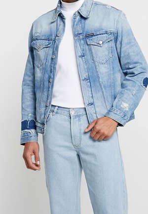 Mann trägt eine hellblaue Jeansjacke mit Used-Details und passende hellblaue Jeans über einem weißen Rollkragenpullover.