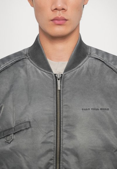 Gray bomber jacket with a zip front, ribbed collar, and a side pocket. Features minimalistic branding and a smooth, matte finish.