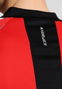 Camiseta atlética roja y negra con un diseño aerodinámico, con un logotipo de la marca prominente impreso verticalmente en la parte posterior.
