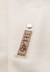 Close-up af et beige stof med en brun firkantet label med teksten "LEVEL UP CN3R" ved siden af en lille rund sølvknap.