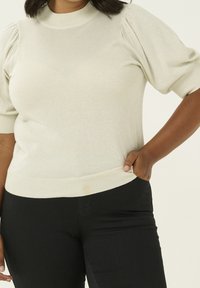 Vrouw in een crèmekleurige korte puff-sleeve trui en zwarte broek, met haar hand in de rechterzak tegen een neutrale achtergrond.