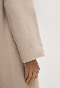 Beige wollen jas mouw met een zachte, gestructureerde afwerking; manchet is licht gerold. Hand die de stof vasthoudt, wat een relaxte pasvorm toont.