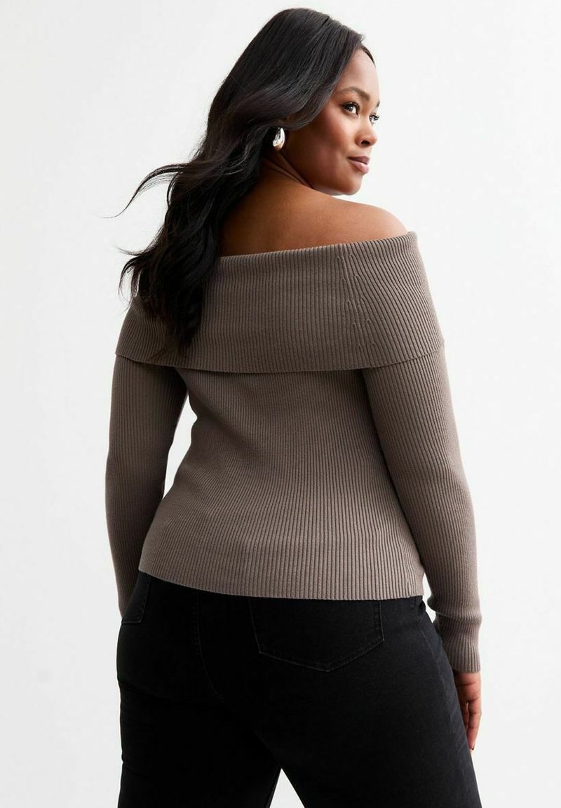Maglione a spalla scoperta in rib ribattuto color taupe con maniche lunghe, presenta un colletto piegato e dettagli testurizzati. Indossato con jeans neri.