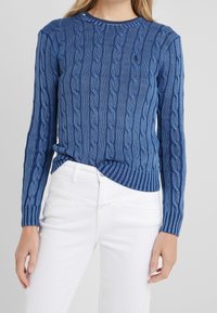 Kvinna iklädd en blå kabelstickad tröja som är nedstoppad i högmidjade vita byxor, stående mot en enfärgad bakgrund.