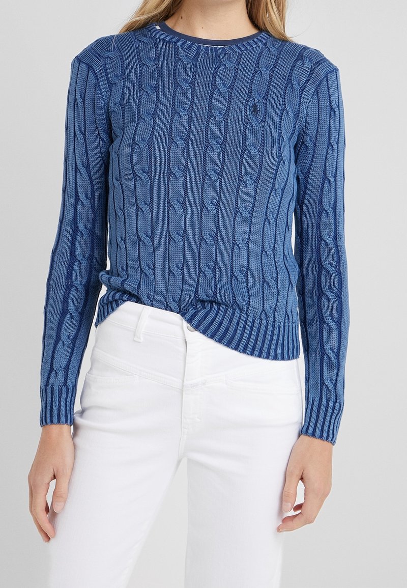 Kvinna iklädd en blå kabelstickad tröja som är nedstoppad i högmidjade vita byxor, stående mot en enfärgad bakgrund.