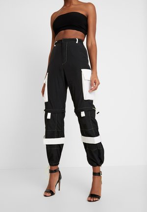Frau trägt schwarzes trägerloses Crop-Top, schwarze Cargohosen mit weißen Taschen und Riemen sowie schwarze Sandalen mit hohen Absätzen vor einfarbigem Hintergrund.