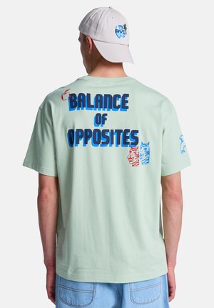 Uomo che indossa una maglietta verde chiaro con scritta blu "Balance of Opposites" e illustrazioni di tigri rosse e blu sulla schiena, cappello bianco e jeans azzurri.
