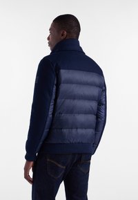 Granatowa kurtka z mieszanką teksturowanej tkaniny i pikowanych paneli, z prążkowanym kołnierzem i mankietami oraz dyskretną marką na rękawie.