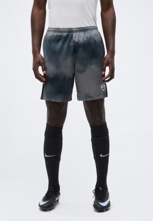 Czarne sportowe spodenki z szarozielonym i ciemnoniebieskim wzorem tie-dye, noszone przez osobę w czarnych skarpetach piłkarskich Nike i kaskach.