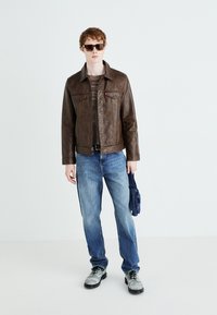 Smeđa kožna jakna s dva džepa na prsima, u paru s plavim trapericama. Model drži tamnoplavu torbu, nosi sunčane naočale i sive cipele.