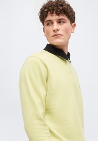 Νεαρός άνδρας με καστανά σγουρά μαλλιά, που φοράει ανοιχτό κίτρινο φούτερ και μαύρο πουκάμισο με γιακά, κοιτάζει πλάγια σε απλό φόντο.