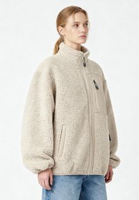 Giacca in pile beige con collo alto, zip frontale, tasche laterali e polsini a coste. Presenta un tessuto strutturato e un design a vestibilità rilassata.