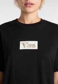 Sort bomuld t-shirt med en rektangulær hvid lap med ordet "Vans" i guldskrift centreret på brystet. Korte ærmer, rund hals.