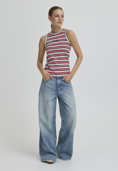 Femme debout les mains dans les poches, portant un haut sans manches rayé rouge, blanc et noir, ainsi que des jeans larges bleu clair, sur un fond uni.