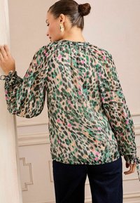 Caura blūze ar pleitu dizainu, kas rotāta ar zaļu, melnu un rozā dzīvnieku rakstu; elastīgas aproces; noapaļots kakla izgriezums; viegla auduma.