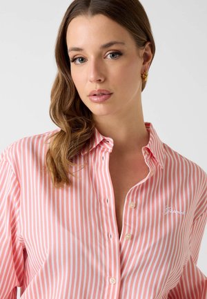 Jeune femme aux cheveux bruns ondulés portant des boucles d'oreilles en or et une chemise à rayures roses et blanches avec un petit logo brodé.