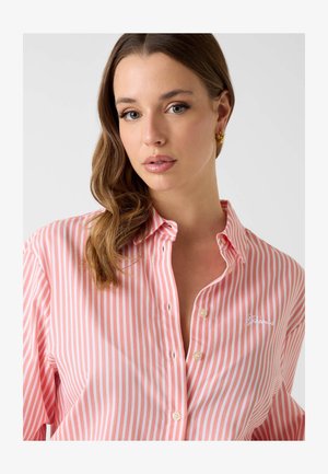 Jeune femme aux cheveux bruns ondulés portant des boucles d'oreilles en or et une chemise à rayures roses et blanches avec un petit logo brodé.