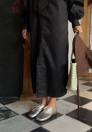 Persoon draagt een zwarte jurk en zilveren veterschoenen, houdt een geweven handtas vast met een geruit doekje erin, en staat op een vloertegel met dambordpatroon.