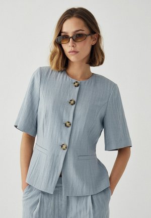 Žena v svetlomodrom krátkomrukávovom saku so zapínaním na gombíky a k tomu ladných nohaviciach, má ruky v vreckách a doplnila vzhľad úzkymi hnedými slnečnými okuliarmi.