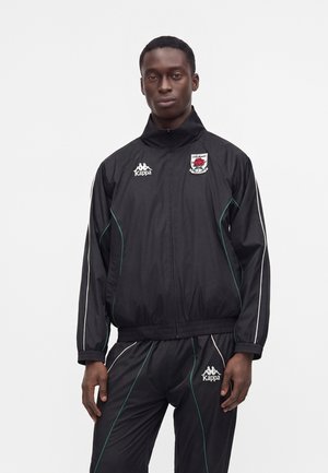 Hombre de pie con un chándal negro de Kappa con detalles en blanco y verde, que muestra logotipos de equipo y de la marca, de frente sobre un fondo liso.