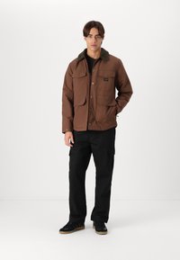 Brun jakke med pelsforet krave, der har flere forlommer, sat sammen med sorte cargobukser og sorte sneakers. Ensfarvet baggrund.