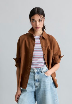Junge Frau mit dunklem Haar, die ein braunes Hemd mit Knöpfen über einem gestreiften Oberteil und hellblauen hoch taillierten Jeans trägt, steht vor einem einfarbigen Hintergrund.
