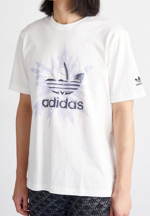 Camiseta de algodón blanca con un logo central de Adidas en negro y gris con un patrón de rayos, mangas cortas y cuello redondo acanalado.