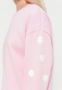 Rosa sweatshirt med avslappnad passform, rundad halsringning och dekorativa vita prästkragar på vänster ärm. Mjuk textur och avslappnad design.