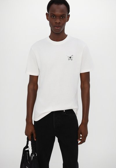 White cotton t-shirt featuring a small black butterfly skull graphic on the left chest. Paired with black denim pants, held with a dark bag.