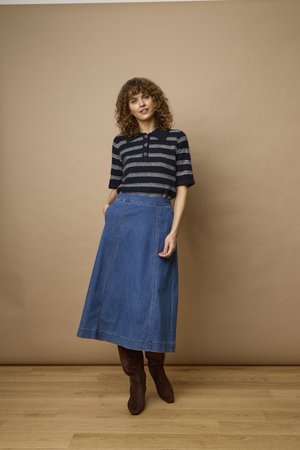 Femme aux cheveux bouclés portant une chemise rayée noir et blanc, une jupe en jean bleue et des bottes marron, debout sur un sol en bois contre un mur beige.