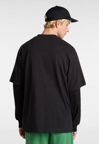 Schwarzes T-Shirt mit kurzen Ärmeln, das über einem langärmligen schwarzen Shirt getragen wird, kombiniert mit grünen Hosen. Hat einen lässigen Schnitt und weichen Stoff.