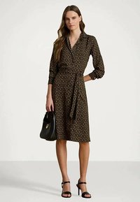 Žena u crnoj i smeškastoj haljini s uzorkom, drži crnu torbicu i nosi crne sandale na visoku petu, stoji pred jednostavnom pozadinom.