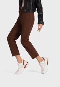 Sneakers bianchi con dettagli marroni, in pelle e mesh, abbinati a pantaloni cropped marroni. L'outfit presenta una giacca in pelle nera elegante.