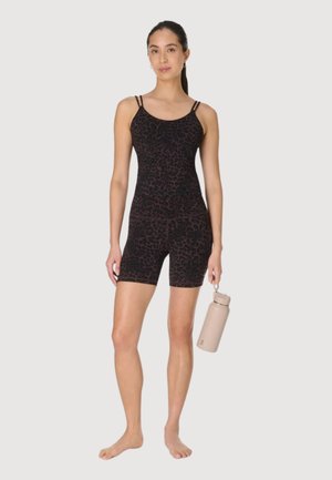Jonge vrouw staat blootsvoets, draagt een donker mouwloos jumpsuit met luipaardprint en houdt een lichtbeige waterfles naast zich vast.