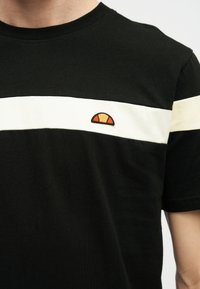 Μαύρο T-shirt με οριζόντια λευκή ρίγα στο ύψος του στήθους και μικρό ημικυκλικό πορτοκαλί και κίτρινο λογότυπο στην αριστερή πλευρά.