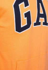 Sudadera naranja con grandes letras en azul marino "GAP" con un contorno blanco; incluye un bolsillo delantero y una tela suave y texturizada.