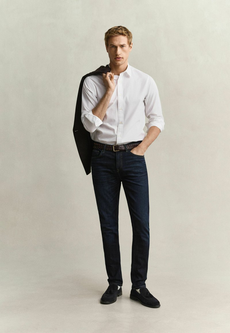 Young man wearing white shirt and dark jeans, holding black jacket over shoulder with one hand and the other hand in pocket.
