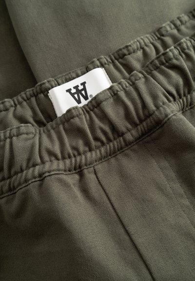 Pantalon taille élastique vert olive avec une étiquette en tissu blanc portant un logo noir cousu à l'intérieur de la ceinture.