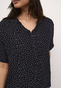 Blusa blu navy a maniche corte con pois bianchi, chiusura a bottoni, scollatura arricciata e morbida texture del tessuto.