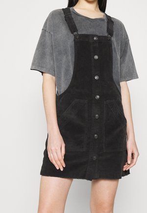 Black corduroy dress with adjustable straps, front buttons, and side pockets. Paired with a faded gray t-shirt underneath.