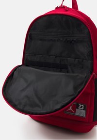 Sac à dos rouge en tissu résistant, doté d'une poche en maille noire zippée, d'une grande ouverture et d'un patch logo avec le numéro 23.