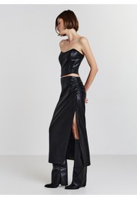 Frau trägt ein schwarzes, trägerloses Crop-Top aus Leder und einen passenden Maxirock mit hohem Schlitz sowie schwarze kniehohe Stiefel, steht seitlich.