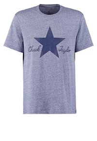 Šedé tričko s krátkým rukávem, které na sobě má tmavě modrou grafiku hvězdy s nápisem "Chuck Taylor" napsaným kurzívou pod hvězdou. Měkká textilie.