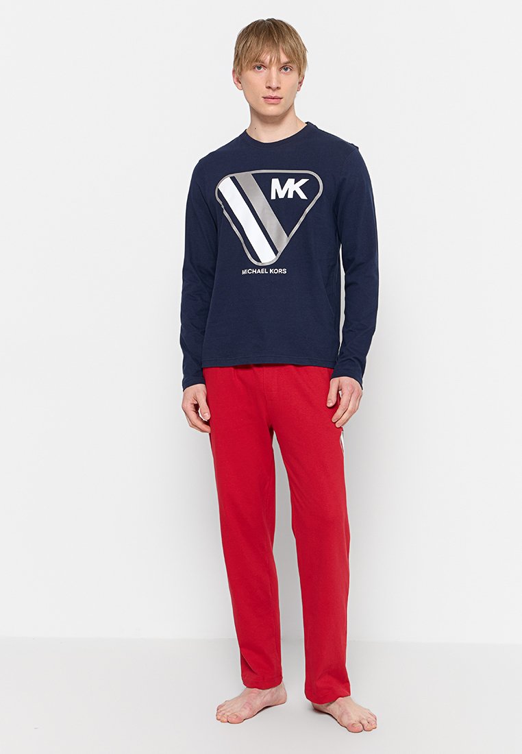 Giovane uomo in piedi a piedi nudi, indossa una camicia a maniche lunghe blu navy di Michael Kors e pantaloni rossi su uno sfondo bianco.