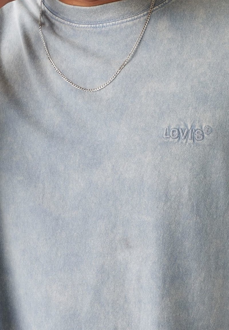 Gros plan d'une personne portant un t-shirt Levi's bleu clair et une fine chaîne en argent autour du cou.