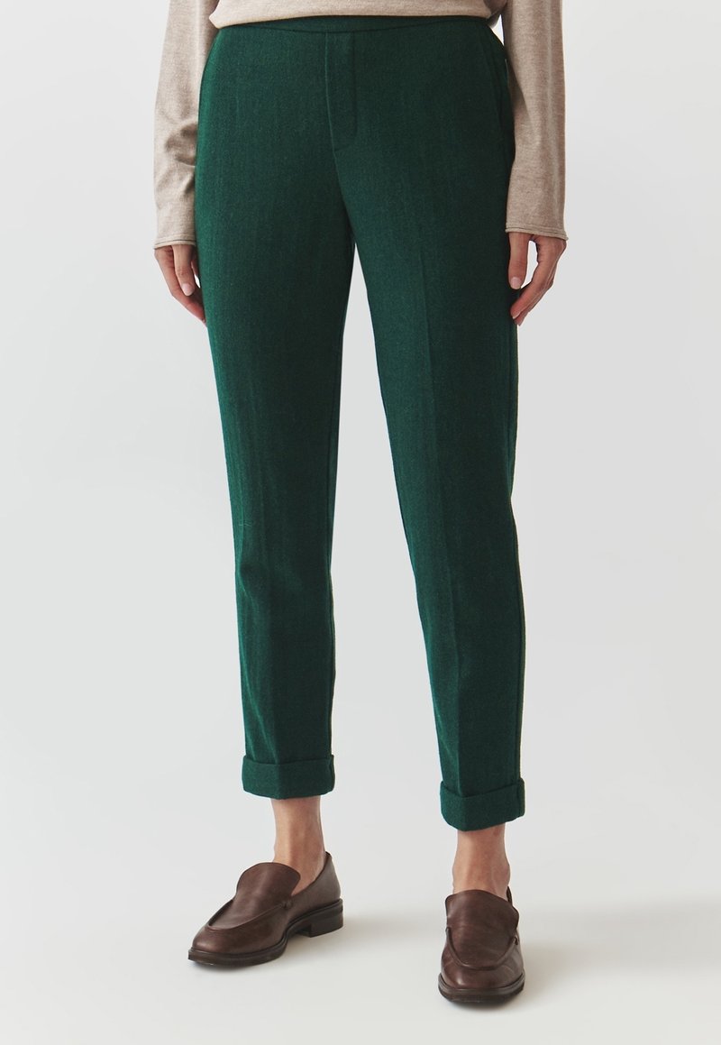 Pantalon vert en laine avec une coupe droite, présentant un pli marqué à l'avant, des ourlets retroussés et des poches latérales. Associé à des mocassins marron.