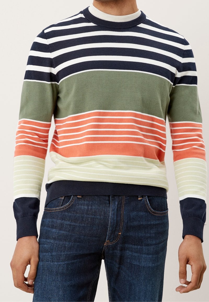 Mann trägt einen mehrfarbigen gestreiften Pullover in Marineblau, Weiß, Grün, Orange und Creme, kombiniert mit dunkelblauen Jeans.