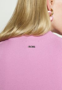 Ribbad rosa tröja med rund halsringning, med en liten svart "BOSS"-logotyp på baksidan. Slät textur och åtsittande design.