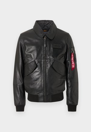 Zwarte leren bomberjack met een ritssluiting aan de voorkant, twee schuine zakken, geribbelde manchetten en zoom, en een rood label met de tekst "Remove Before Flight" op de zak op de linkermouw.